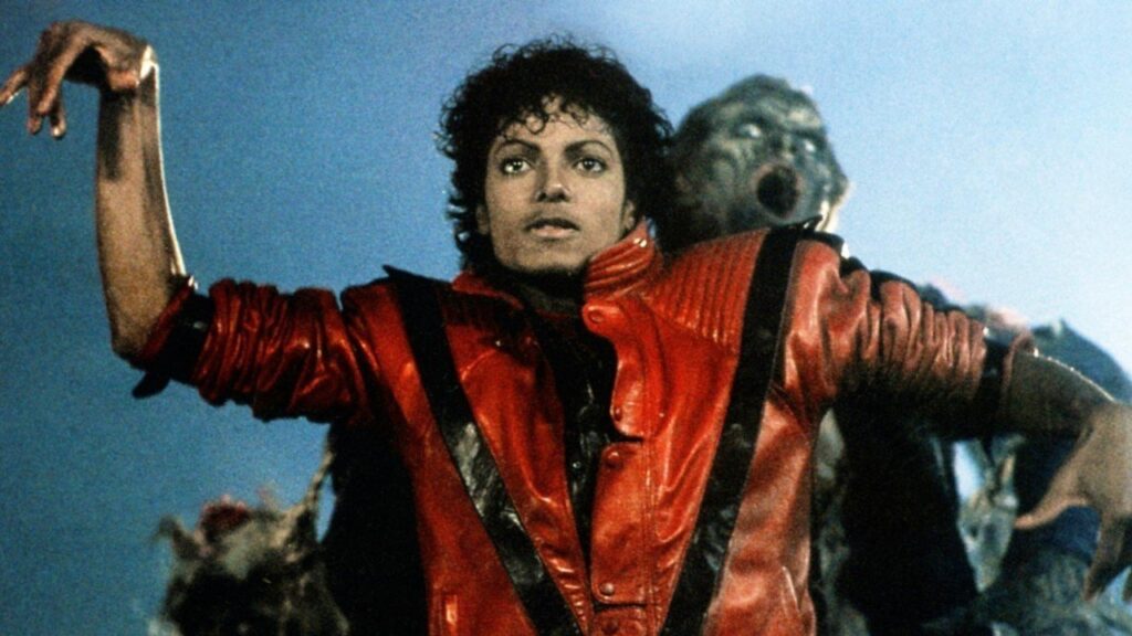 Promotional still from Michael Jackson’s “Thriller” music video featuring Jackson in his iconic red leather jacket, surrounded by zombie dancers in a dimly lit urban setting. The image captures the fusion of theatrical fashion and pop culture that redefined music video aesthetics and streetwear influence in the 1980s.