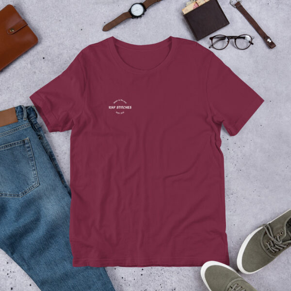 A mahroon tshirt with the logo rapstitches and objects like jeans, glasses, shoes and wallet in the background
