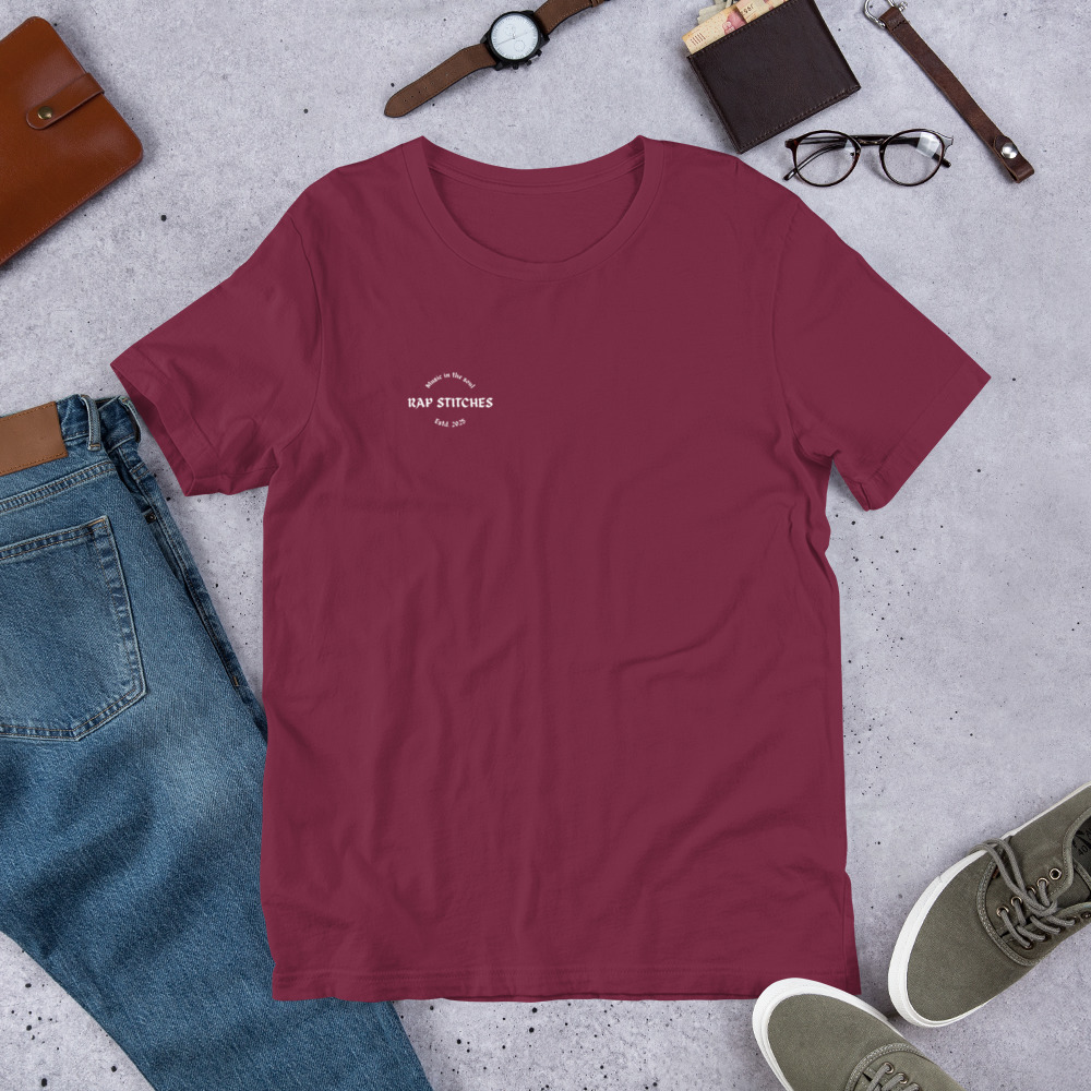 A mahroon tshirt with the logo rapstitches and objects like jeans, glasses, shoes and wallet in the background
